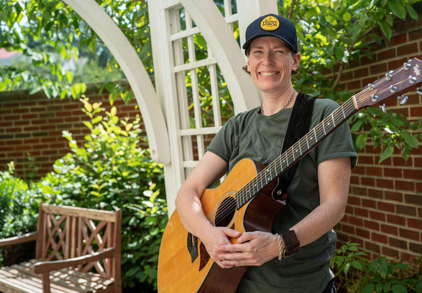 Clara, Front Porch Music volunteer wearing a green shirt and blue Front Porch hat holding a guitar and smiling on a front porch.