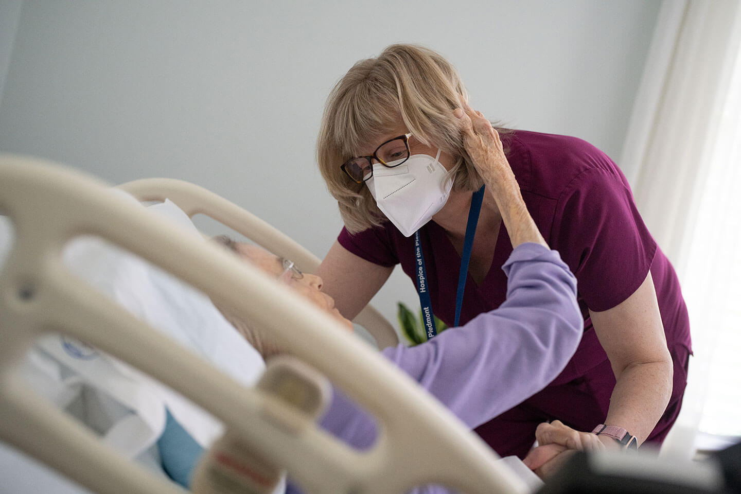 Hospice nurse wearing a mask reaching down to a patient in her bed who is wrapping her arm around the nurse's neck