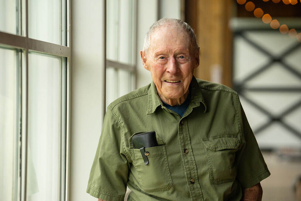 An elderly hospice patient in a green shirt standing by a window and smiling.