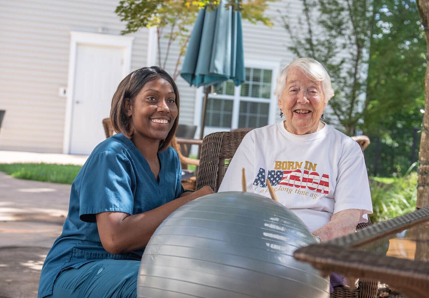 A hospice patient and nurse smiling and playing the drums on an exercise ball while sitting at a picnic table.