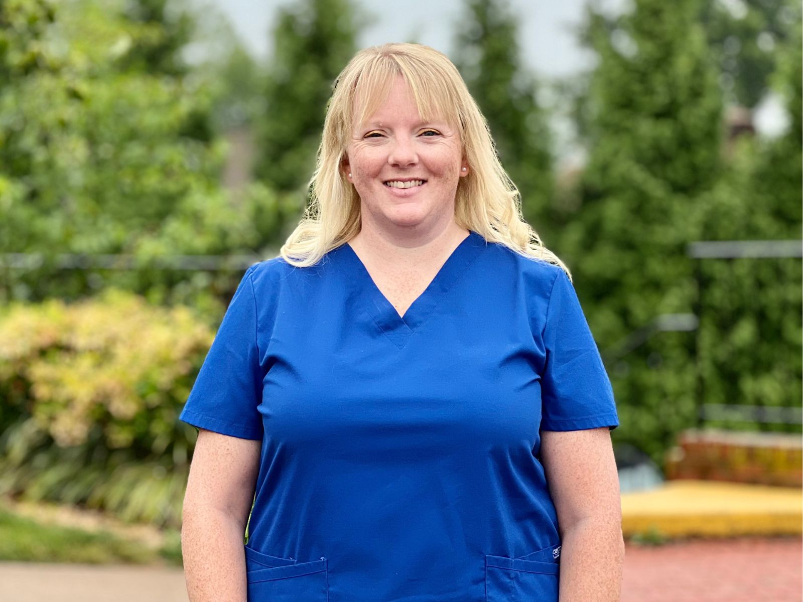 Katy Strain in blue scrubs smiling in front of blurred out trees and sidewalk