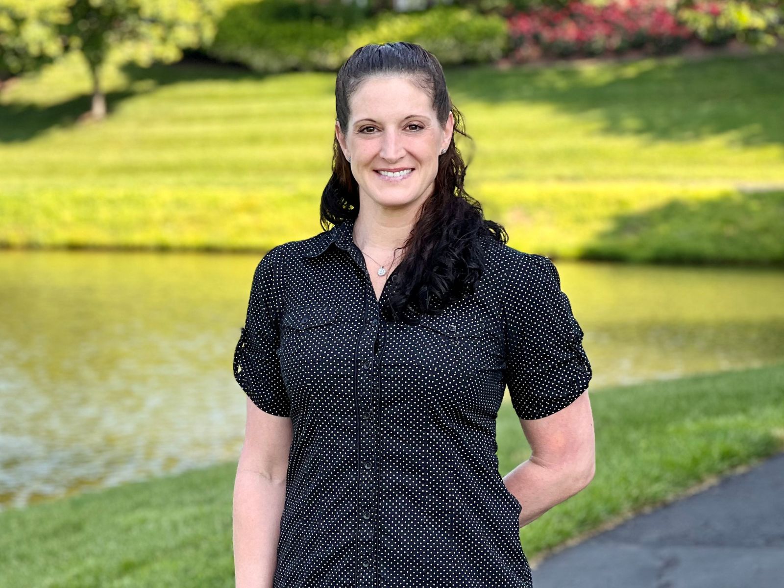 Suzette Doran smiling and standing in front of a pond with grass and a sidewalk.