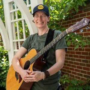 Musician Clara George wears a green T-shirt and trucker cap with "The Front Porch" logo. She holds an acoustic guitar.
