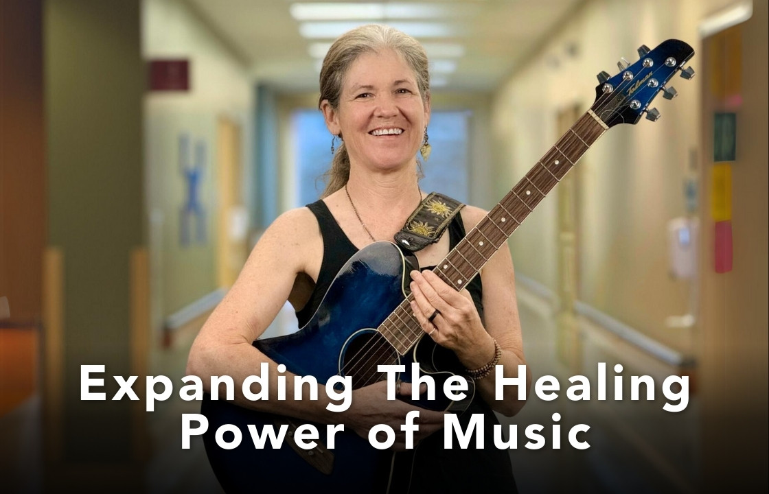 Mary Shapiro plays supportive music at hospice of the piedmont. She holds an acoustic guitar with a blue body. She wears a black tank top.