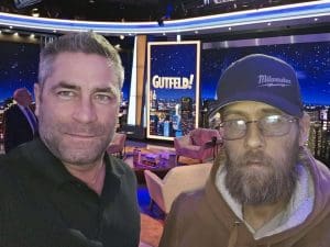 A man in a black button down shirt and a bearded man in a baseball hat and brown hoody stand on the set of the TV show Gutfeld!