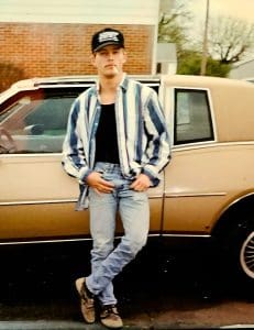 A young man with a mustache and cigarette stands in front of a gold car. He wears a baseball cap, black t-shirt, stripes button down shirt and faded blue jeans.