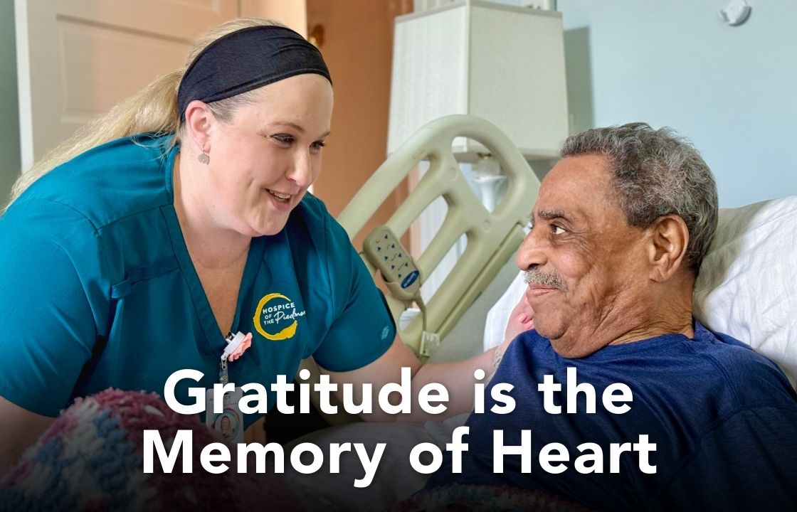 Ashley a hospice of the Piedmont LPN wears teal scrubs and speaks with a patient in a bed at the Hospice House. The words "Gratitude is the Memory of the Heart" appear below them.