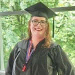 Hospice of the Piedmont Nurse Jessica wears a black graduation cap and gown. She smiles from inside a gazebo in front of leafy trees