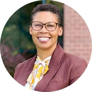 Kimberlyn McDonald wears a desaturated dark red blazer, a yellow and white floral shirt with tied at the top, and glasses.