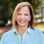 Hospice of the Piedmont CEO wears a light green linen button down shirt. We see a little nature behind her.