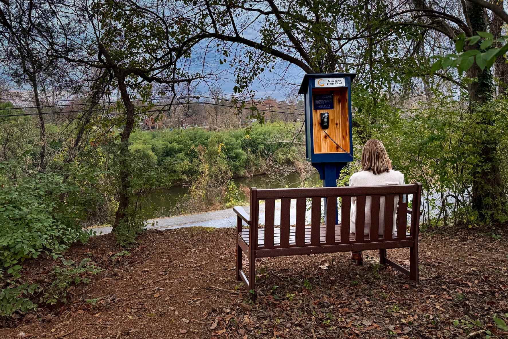 A woman with blond hair sits on a brown bench in the woods. She holds a black phone to her ear, connected to a phone box labeled "Hospice of the Piedmont Telephone of the Wind."