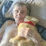 An older man lies in a hospital bed with his robotic companion pet - a golden retriever puppy with a red bandana