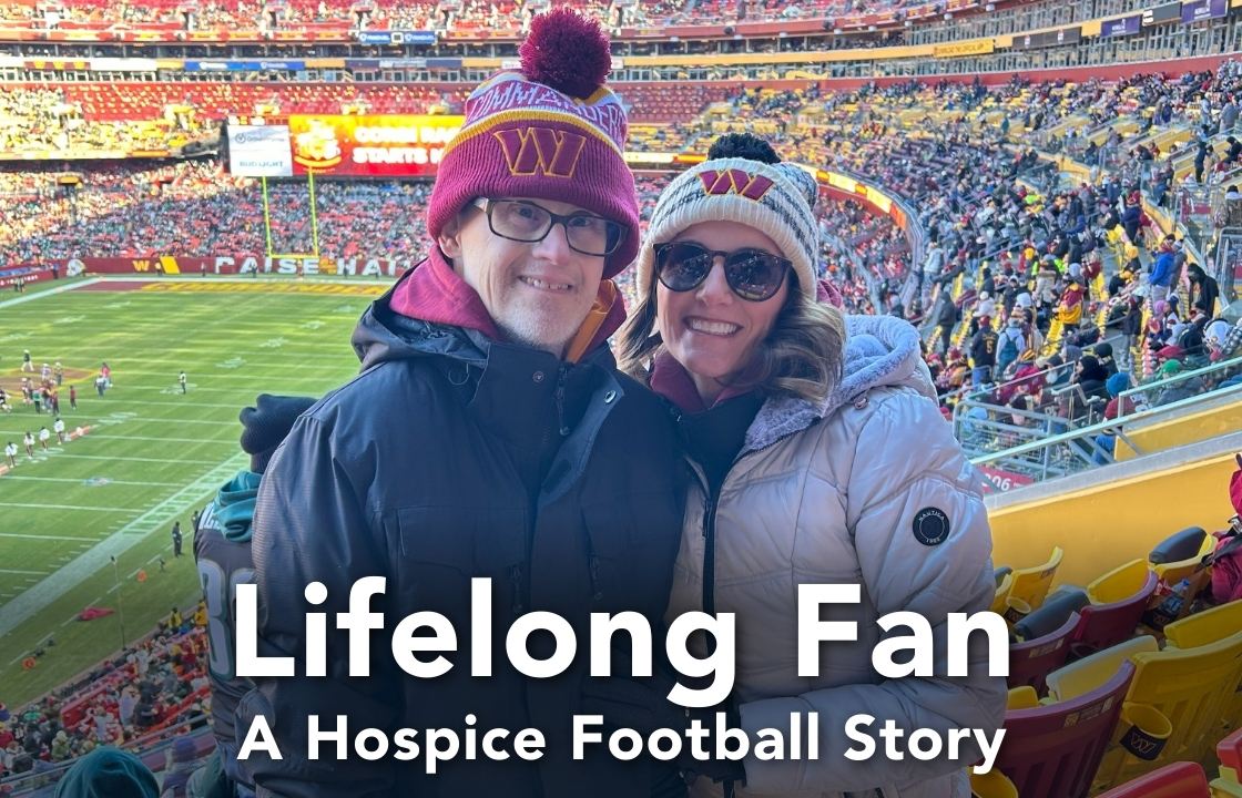 A Hospice patient in a Washington Commanders beanie and black coat, stands alongside his niece also in a Commanders beanie, and white coat. Behind them the Commanders stadium is partially full while teams warm up on the field.