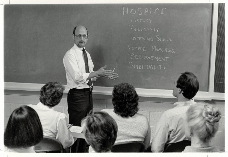 Black and white photo of a man with glasses and wearing a tie standing in front of a blackboard teaching about hospice. In the foreground, a group of students sit at desks; we see only the backs of their heads.