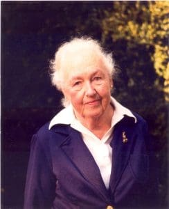 An older woman with thins white hair, wears a purple blazer and wide collared white blouse