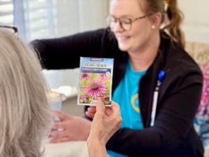 In the foreground, we see the back of a woman's head. Her hair is greying and she holds a seed packet with a bright purple flower. Beyond her and slighly out of focus, a blonde nurse in a bright teal t-shirt is working with potting soil.
