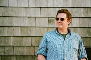 a young man in a light blue button down shirt wears sunglasses in front of a shingled wall. He gazes into the distance.