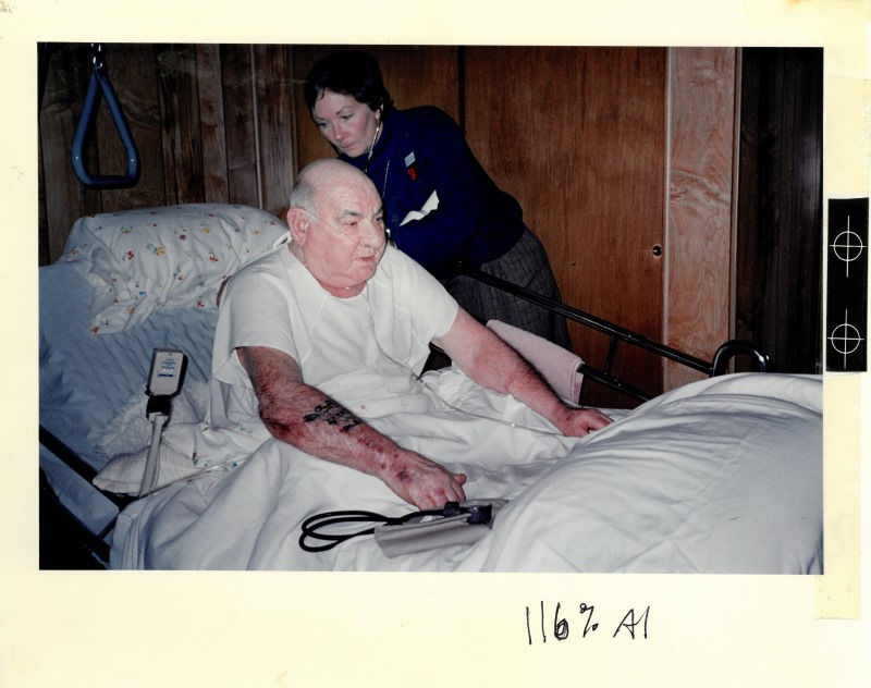 An older man, balding, in a whit t-shirt with a tattoo on his right forearm sits up in a hospital bed while a nurse with short brown hair uses a stethoscope to listen to his back.
