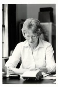 Black and white photo of a Woman Working at Desk with Rolodex. She has large glasses, shorter hair and holds a white piece of paper