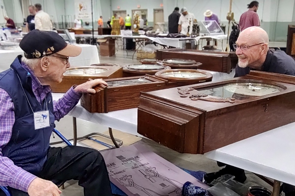 A hospice patient in a wheel chair and baseball hat chats with a goateed smiling man. Between them are a table of wooden mechanical clocks.