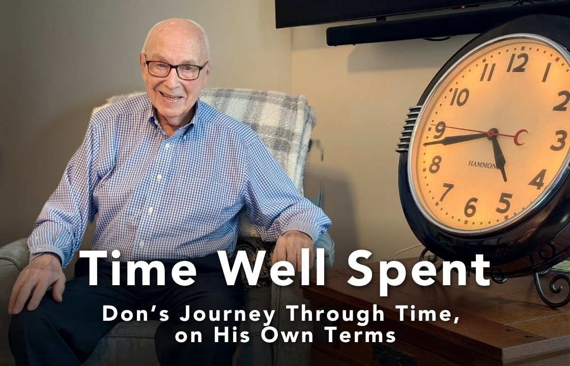 An older gentleman with classes and a button down shirt sits in a comfortable chair next to a large, art deco Hammond clock.