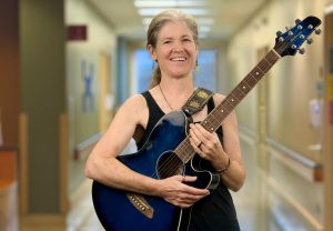 Smiling hospice music therapist holding an acoustic guitar in a softly lit healthcare hallway, conveying warmth, comfort, and healing through music.