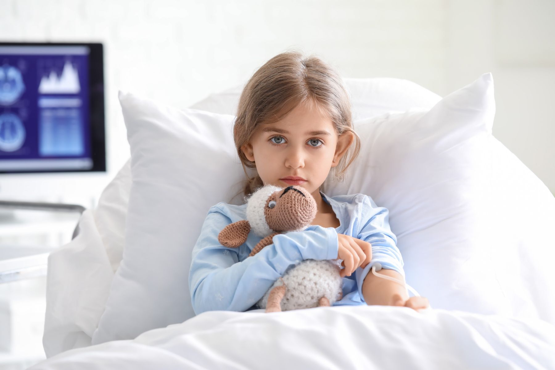 Child sitting upright in a hospital bed for pediatric care holding a stuffed animal, with a medical monitor visible in the background.