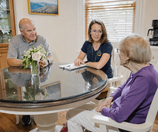 Hospice care team member meets with two older adults around a dining table in a home, discussing care and support in a comfortable kitchen setting.